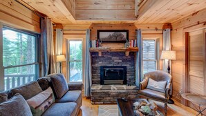 A cozy cabin interior with a stone fireplace, wooden walls, a leather sofa, and a dining table with a rustic centerpiece. - Heritage Hill Cabins (Banner Elk)