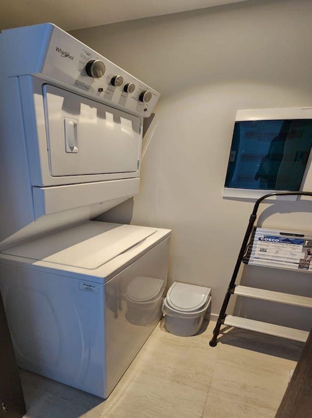 A compact laundry room with a Whirlpool washer and dryer, a trash bin, and a small window.