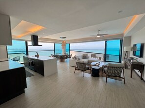 A modern living space with a kitchen, dining area, and seating by a large window overlooking the sea.