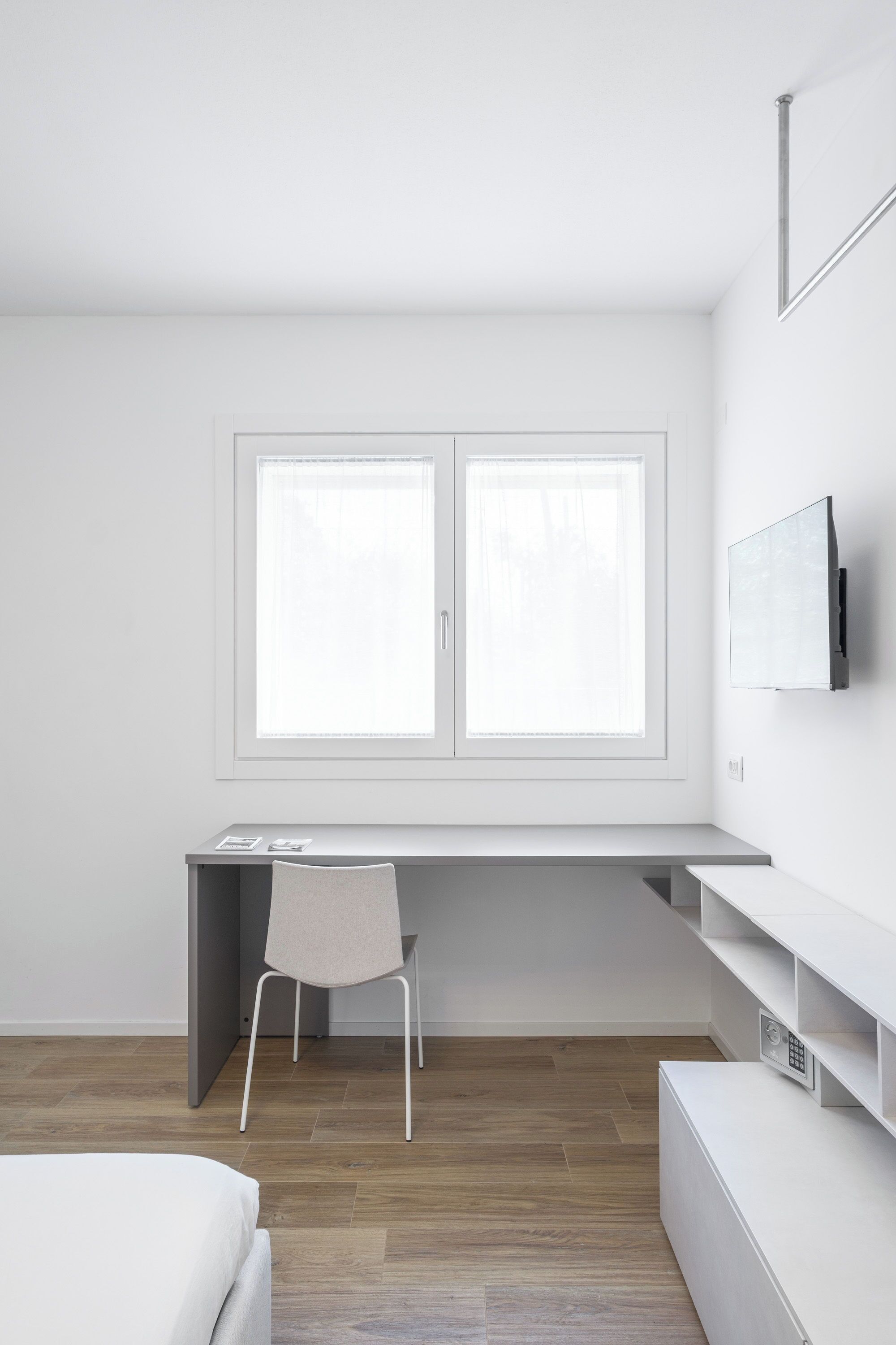 A minimalist bedroom with a bed, a desk with a chair, a TV mounted on the wall, and a window with blinds.
