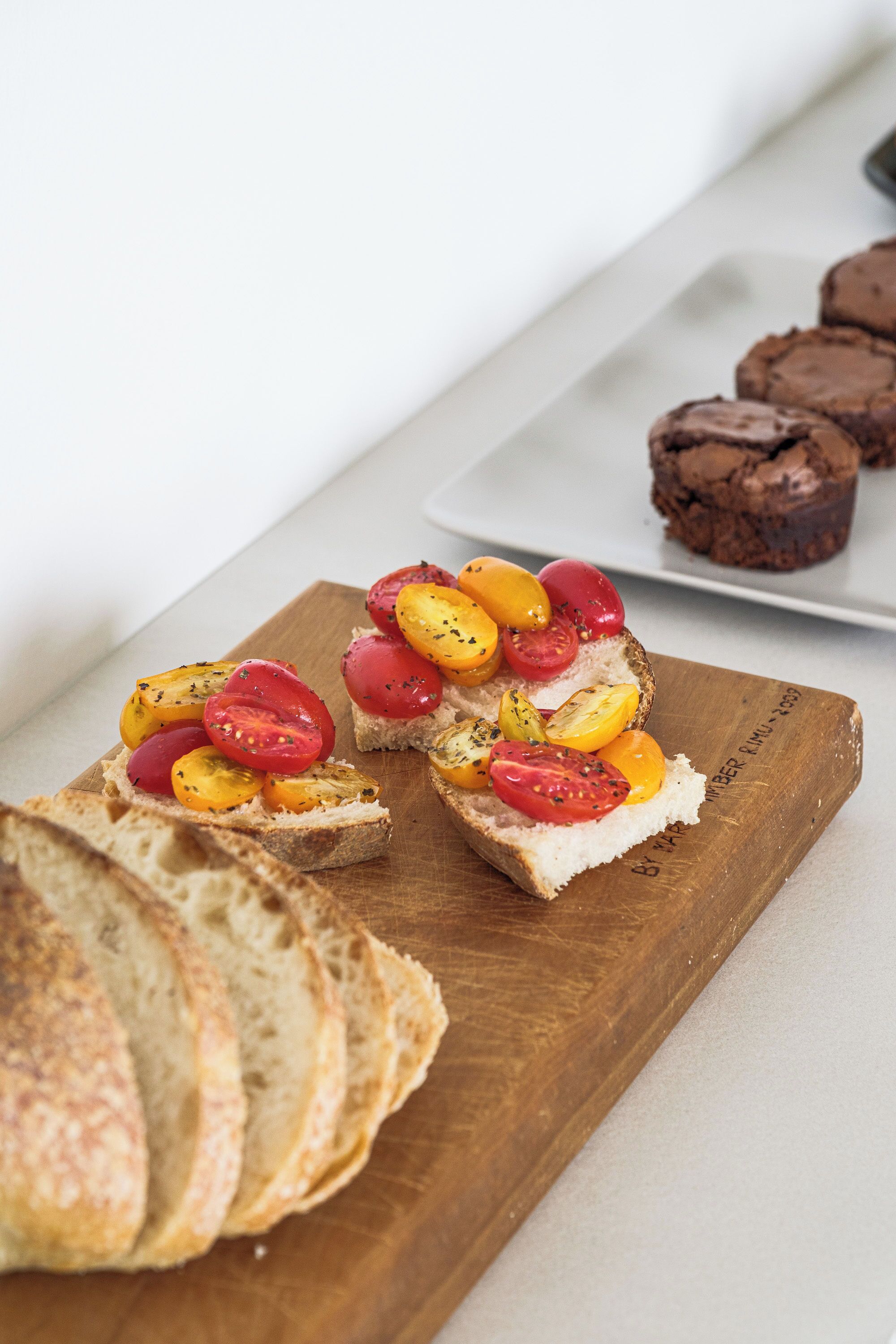 A wooden board with sliced bread topped with tomatoes and herbs, and a plate with chocolate muffins in the background.