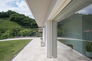 Une terrasse extérieure moderne, dotée d’un mur en verre, d’un sol carrelé et offrant une vue sur une colline verdoyante.