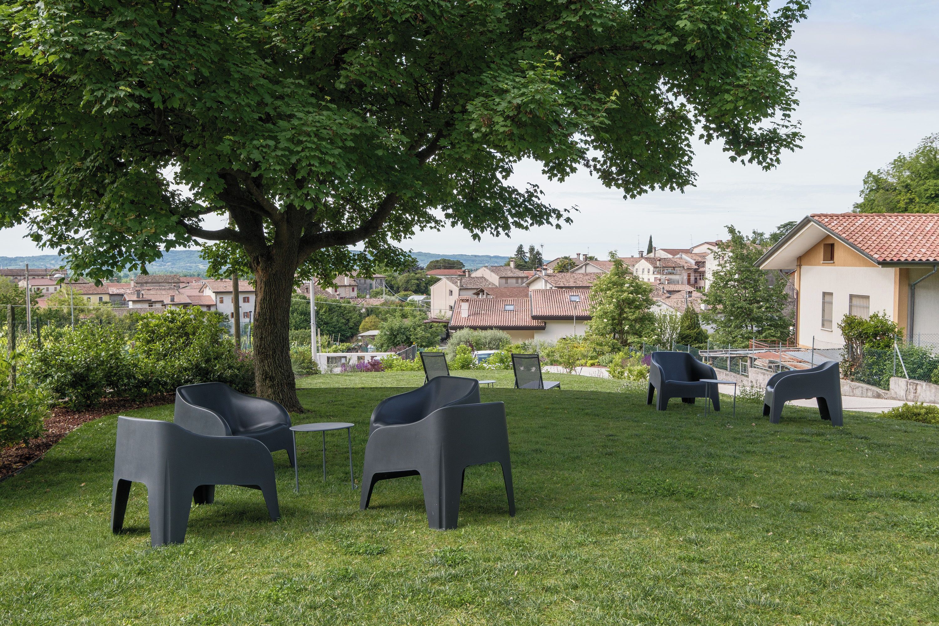 Un espacio al aire libre con césped, sillas modernas y un árbol, con vistas a un barrio residencial.