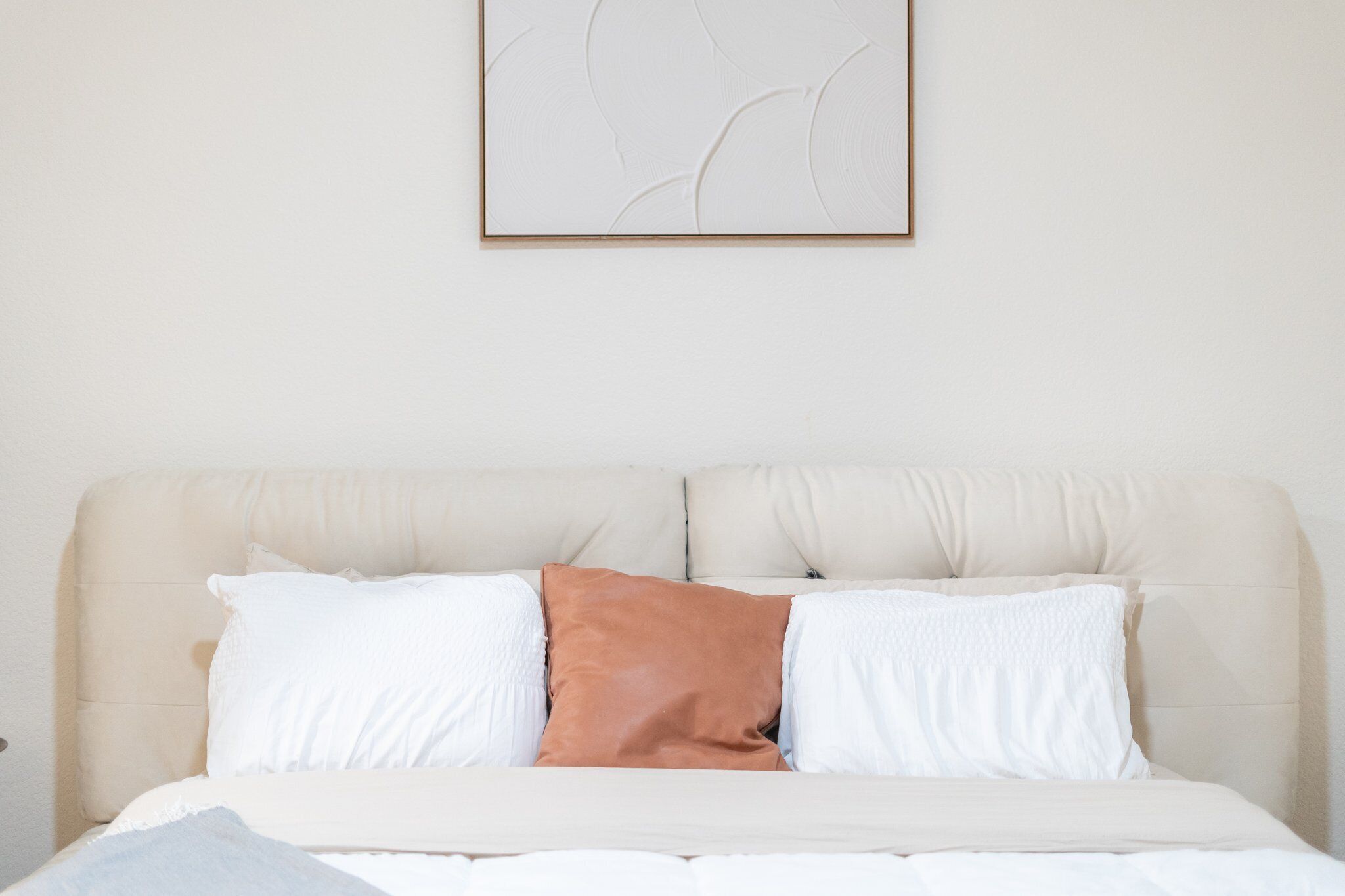 Ein Bett mit einem beigen Kopfteil, weißen und braunen Kissen und einer schlichten Wand mit einem gerahmten Kunstwerk darüber.