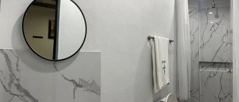 A bathroom with a marble backsplash, a round mirror, a towel rack, and a shower with a marble design.