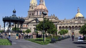 Un edificio histórico con una cúpula destacada y agujas, rodeado por una plaza con bancos y un pabellón.