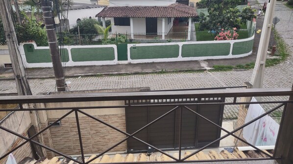 A balcony with a metal railing, a closed shutter door, and a view of a street with a paved road and a building with a tiled roof.