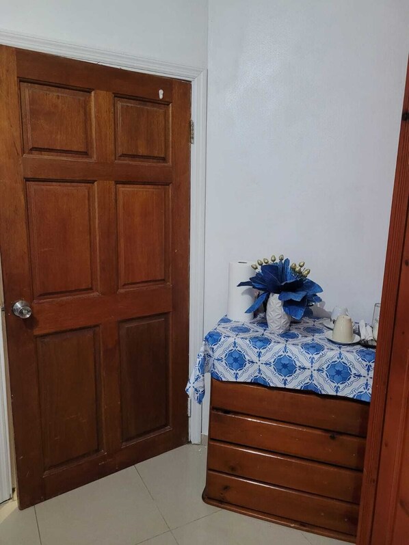 A room with a wooden door, a small table with a blue and white patterned tablecloth, and a white vase with blue flowers.