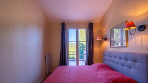 Une chambre à coucher avec une couvre-lit rouge, une tête de lit rembourrée de couleur grise et une fenêtre avec des rideaux.