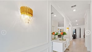Un pasillo con pared blanca, un espejo con marco dorado y una consola blanca con un florero.