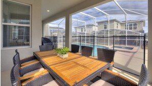 Un patio techado con una mesa de madera y sillas de mimbre, con vista a una piscina y casas vecinas.
