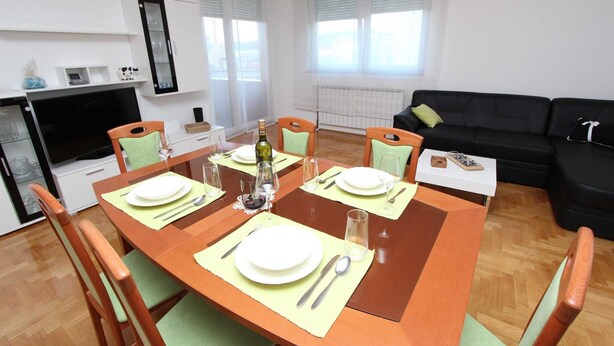 Zona pranzo con tavolo in legno apparecchiato per quattro persone, circondato da sedie. Si notano un divano nero e un armadietto bianco sullo sfondo.