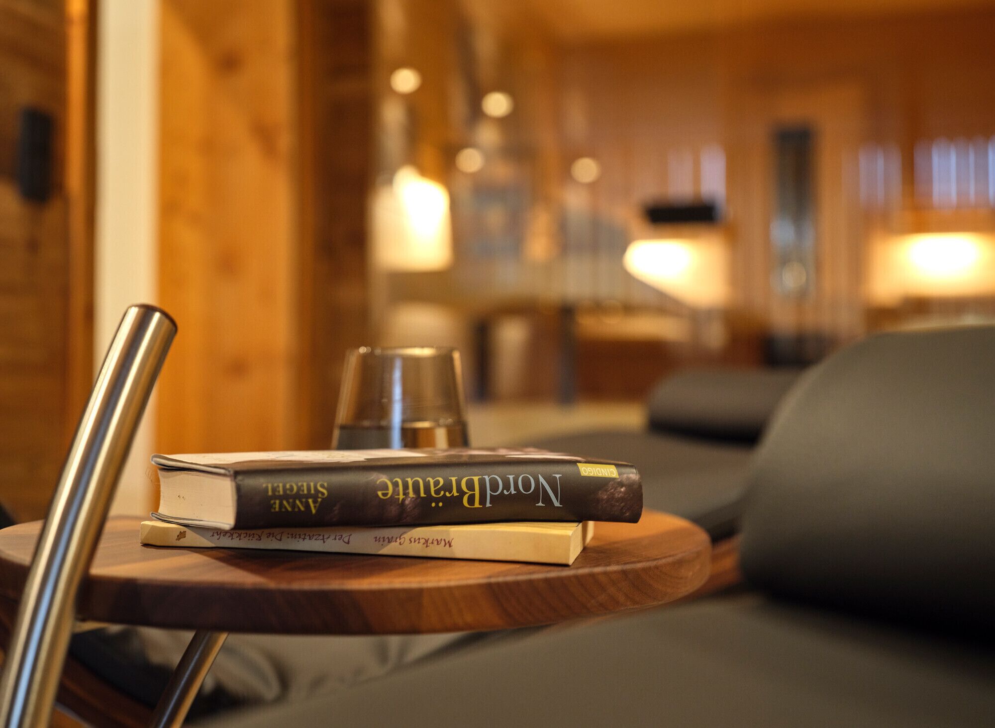 A wooden table with books and a glass on it, a lamp in the background, and a sofa.