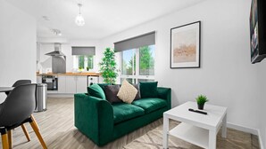 Een moderne woonkamer met een groene bank, een witte salontafel en een kleine plant. Achtergrond is een keuken met een groot raam.