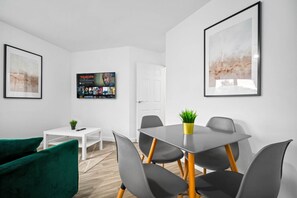A modern room with a dining table, chairs, a green sofa, a small white table with a plant, and two framed artworks on the walls.