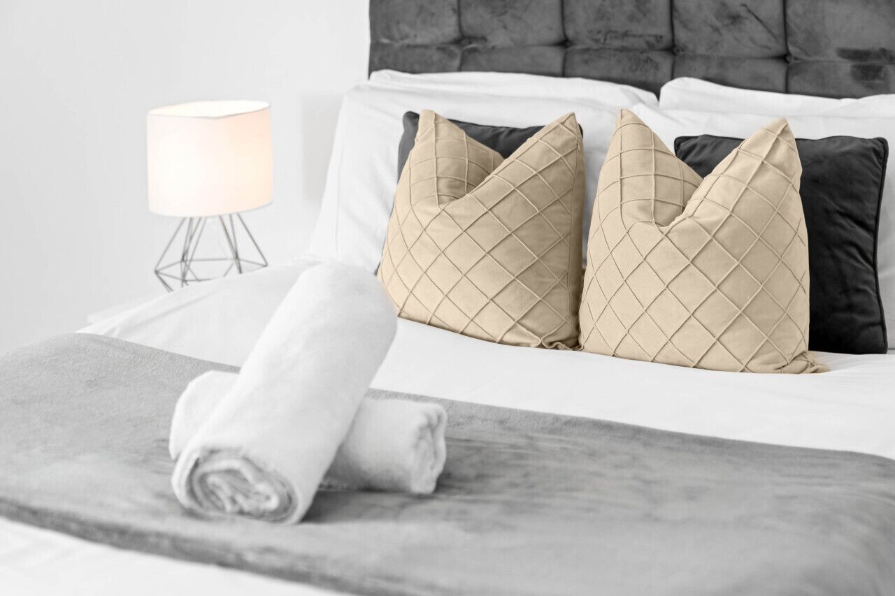 Una cama bien hecha con una toalla enrollada, dos almohadas beige con estampado de rombos, una almohada negra y una lámpara de noche blanca sobre un soporte de metal.