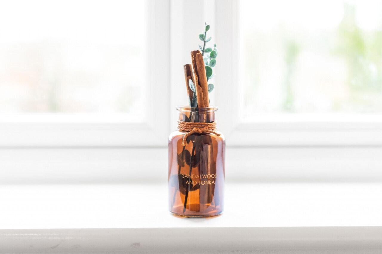 Eine Glasflasche mit Zimtstangen und einer Pflanze auf einem Fensterbrett. Auf dem Etikett ist 'Sandelholz und Tonka' zu lesen.