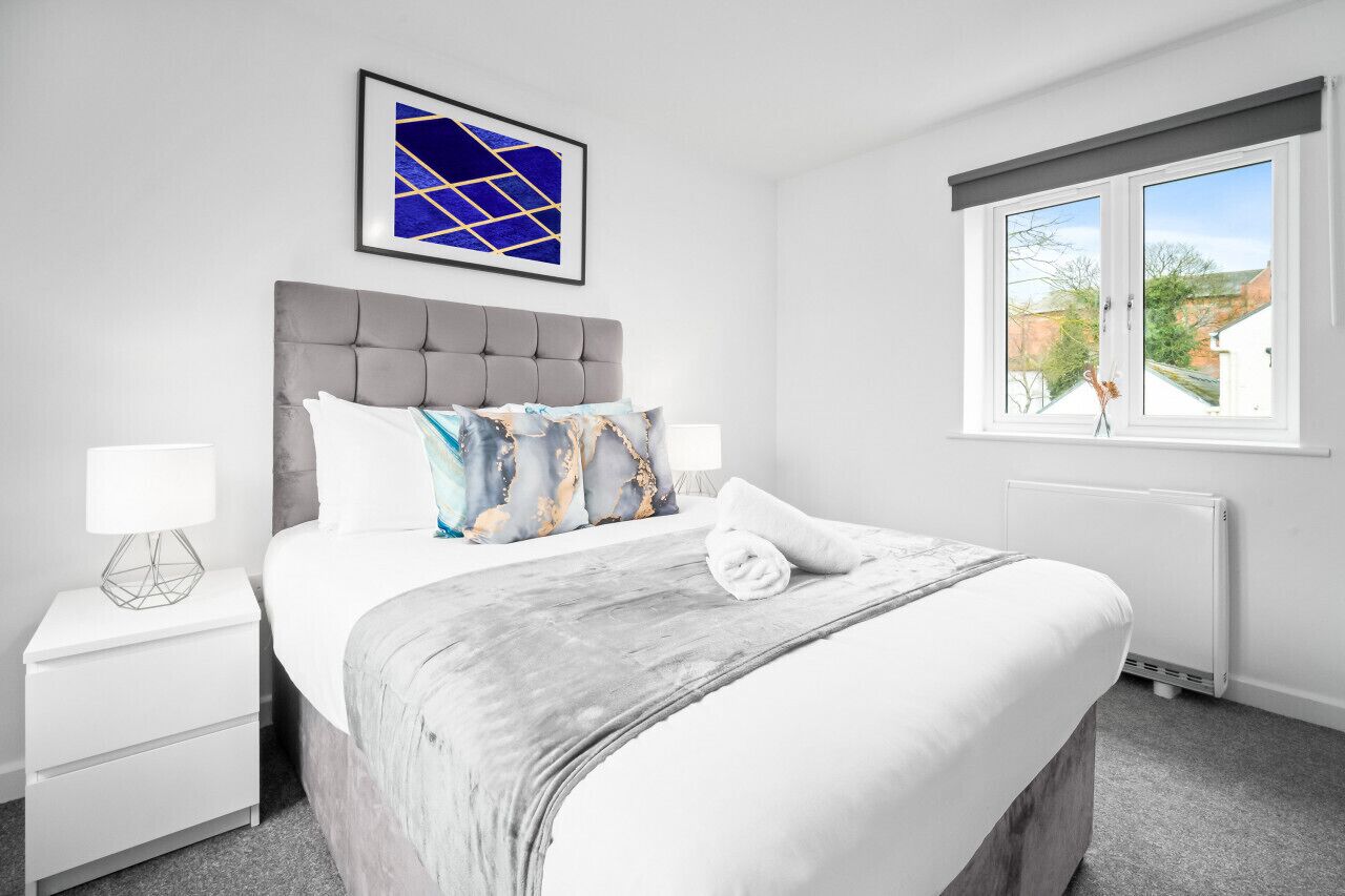 Een moderne slaapkamer met een grijs bekleden hoofdeinde, witte beddengoed en een blauw abstract schilderij aan de muur.
