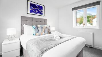 Een moderne slaapkamer met een grijs bekleden hoofdeinde, witte beddengoed en een blauw abstract schilderij aan de muur.