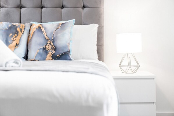 Una cama bien hecha con edredón blanco y dos almohadas, una con diseño de mármol. Una mesita de noche blanca con una lámpara geométrica está al lado de la cama.