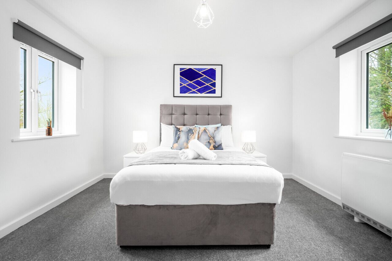 Ein modernes Schlafzimmer mit einem grau gepolsterten Bett, weißer Bettwäsche, einem blauen abstrakten Kunstwerk über dem Bett und einem grauen Teppich.