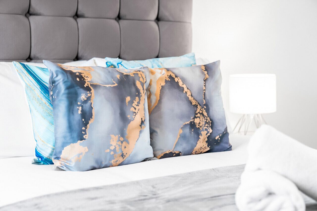 A bed with a grey upholstered headboard, two decorative pillows with a blue and gold abstract pattern, and a white bedside lamp.