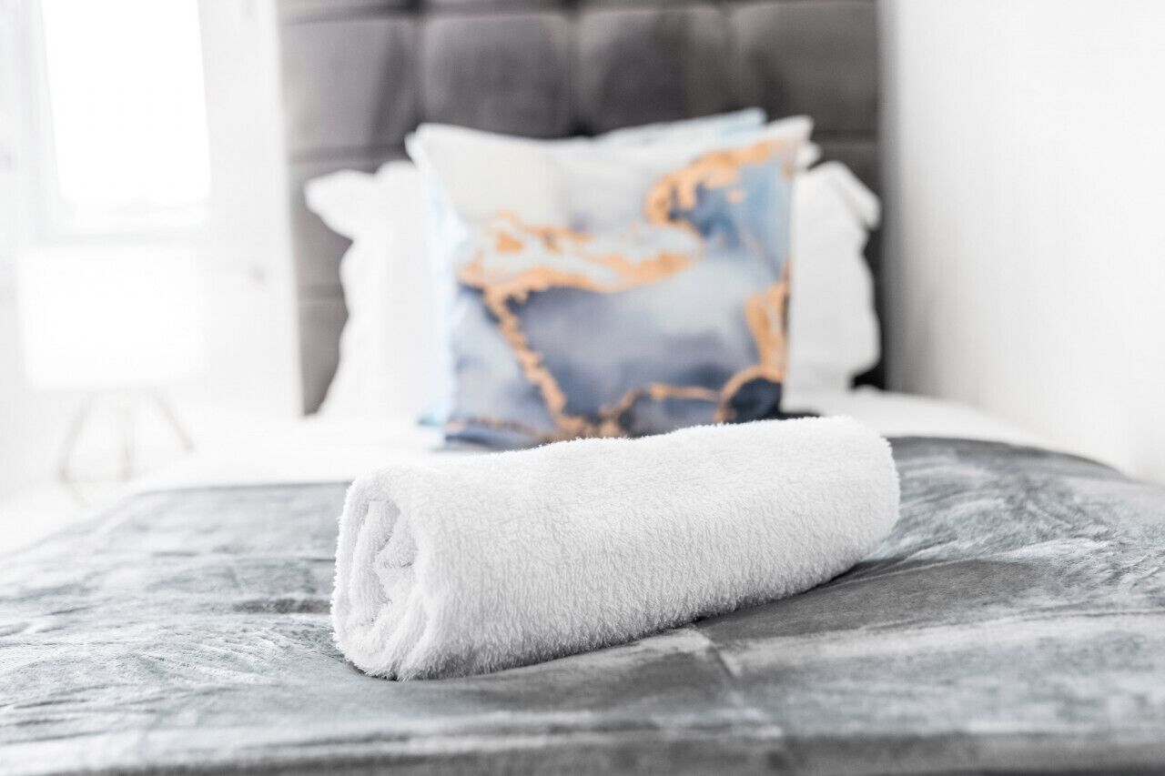 Een netjes opgemaakt bed met een opgerolde witte handdoek en een decoratief kussen met een blauw en goud patroon.