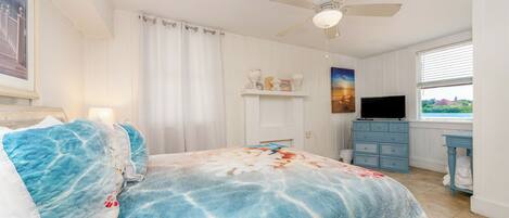 Un dormitorio con cama, ventilador de techo, televisor, cómoda y ventana con vista.
