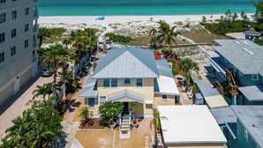Uma propriedade à beira-mar com uma casa central com telhado inclinado, rodeada por uma piscina, palmeiras e uma praia de areia.