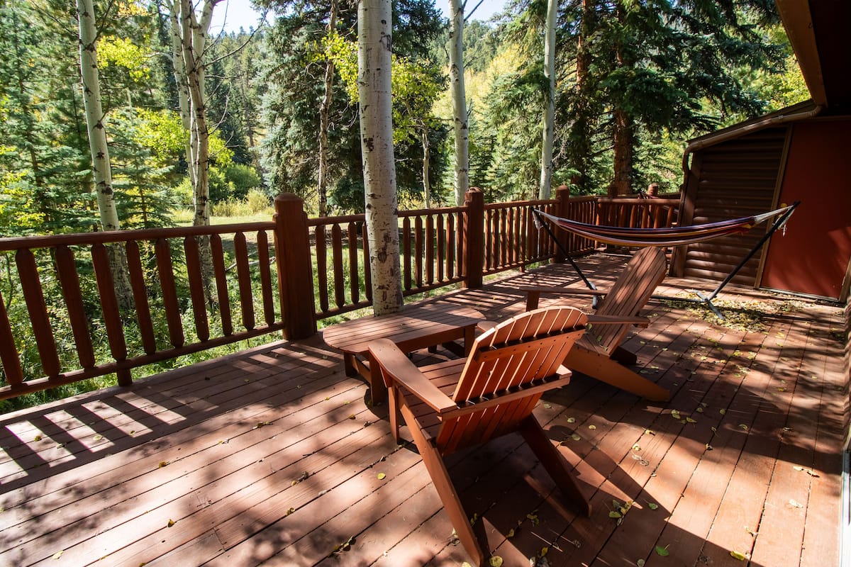A wooden deck with a hammock, chairs, and a table, surrounded by a forest.