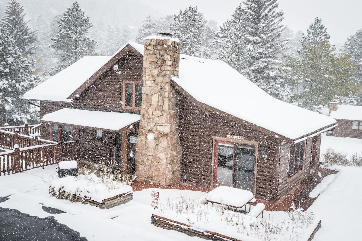 A log cabin with a stone chimney, surrounded by snow-covered trees.