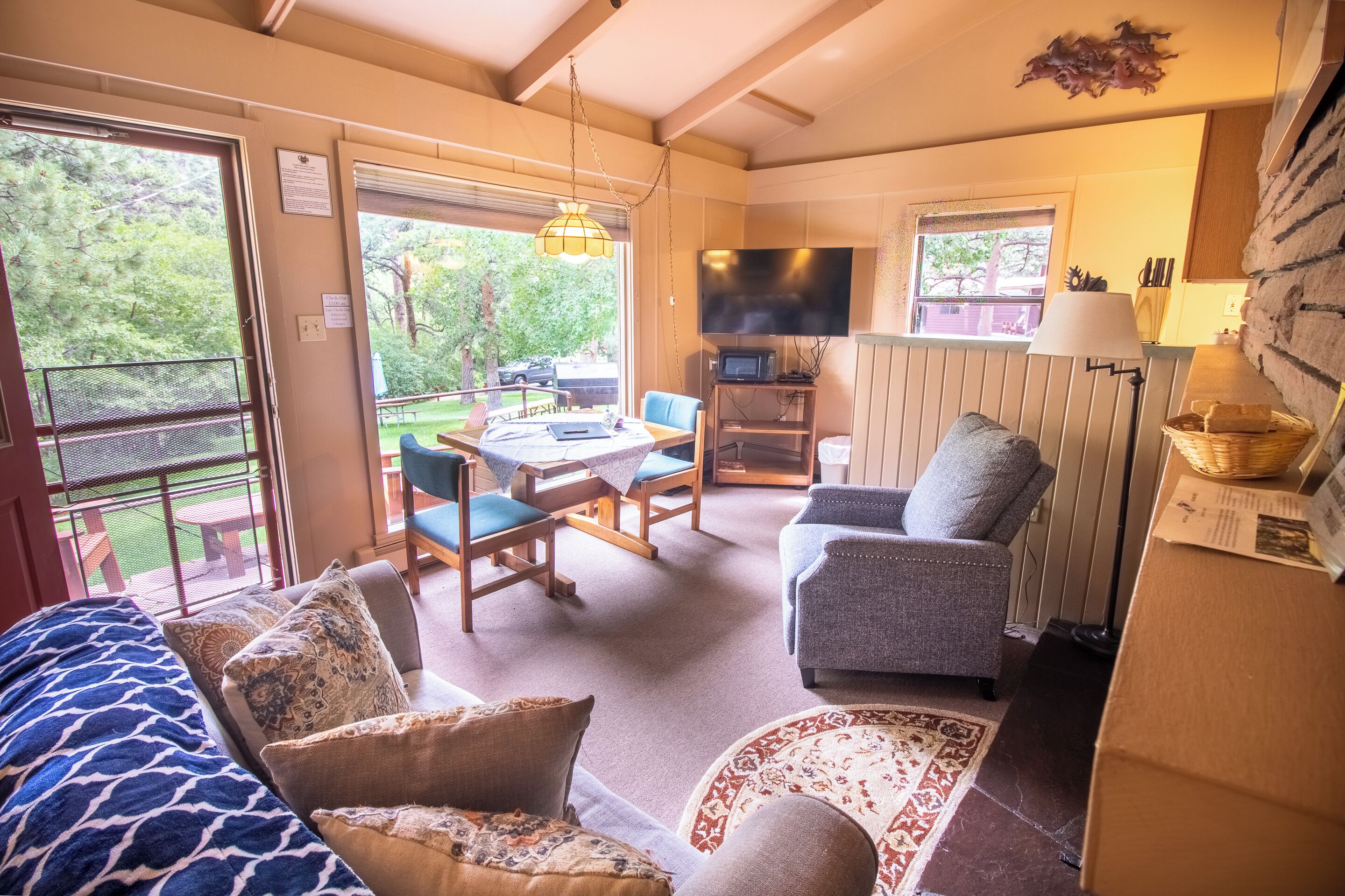 A cozy living room with a sofa, armchair, and a dining table with chairs. There is a TV mounted on the wall and a lamp on a side table.