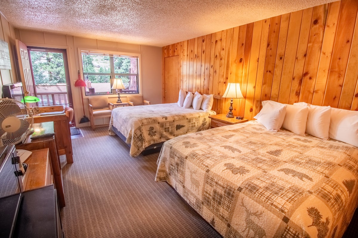 A room with two beds, a desk with a lamp, a chair, and a window with a view of trees.