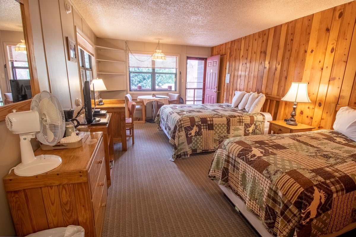 A cozy room with two beds, a desk with a chair, a fan, and a window with blinds.