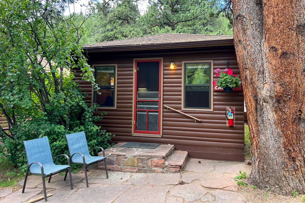 A small cabin with a red door, two windows, a fire extinguisher, and a bench outside.