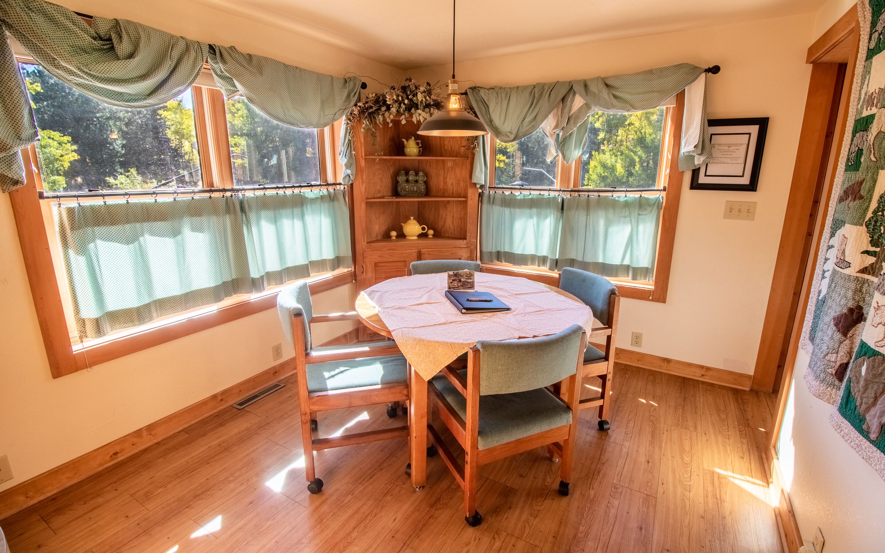 A corner dining area with a round table, two chairs, a wooden cabinet with a shelf, and a window with curtains.