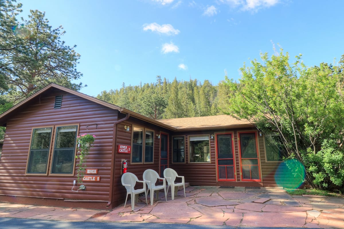 A wooden cabin with a sign reading "River Castle 5," a fire extinguisher, and two white plastic chairs outside.