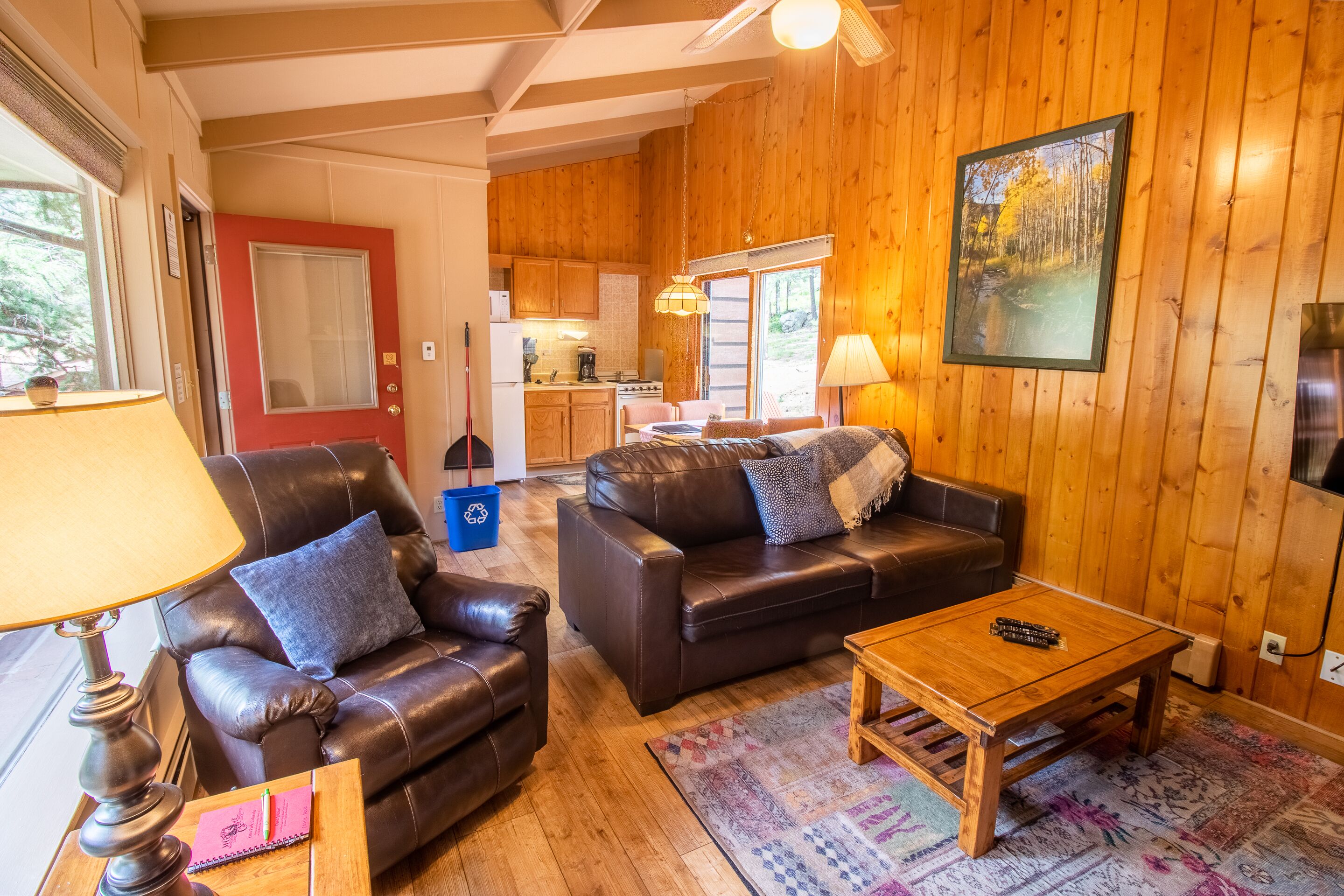 Ein gemütliches Wohnzimmer mit Ledersofas, einem Holztisch und einem Teppich. Im Hintergrund ist eine Küche mit Holzwänden zu sehen.