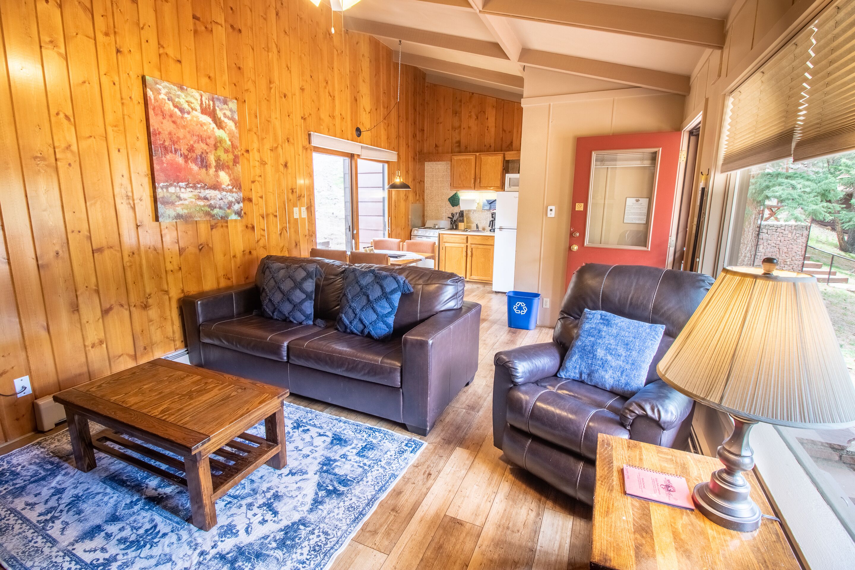 A cozy living room with a wooden floor, a brown leather sofa, a wooden coffee table, and a blue patterned rug.