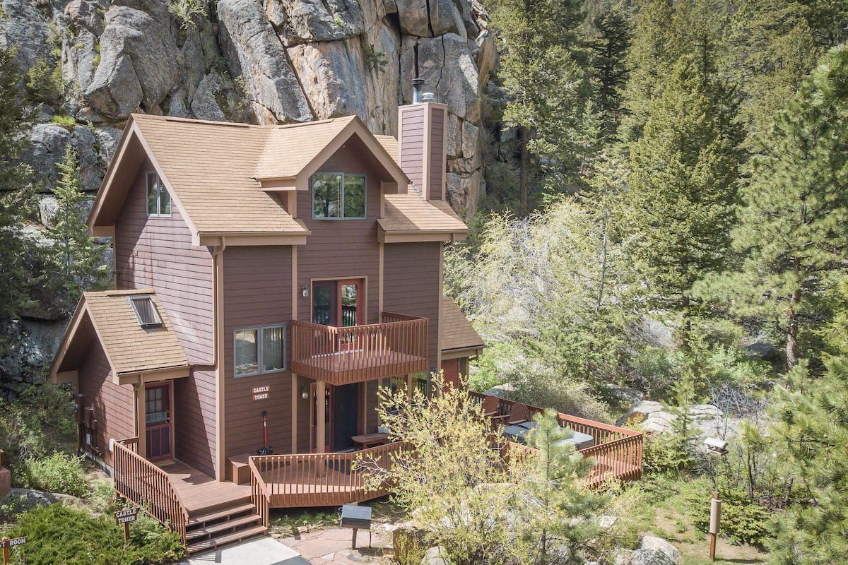 A multi-story cabin with a balcony, surrounded by forest and rocky terrain.