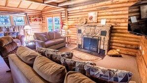 A log cabin interior with a stone fireplace, wooden walls, and a cozy seating area with a sofa and armchairs. - Castle Mountain Lodge (Estes Park)