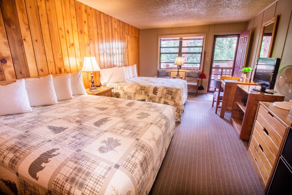 A room with two beds, wooden walls, a desk, a chair, a fan, and a dresser.