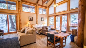 A cozy cabin interior with a wooden floor, a sofa, a chair, and a bed. Large windows offer views of a snowy landscape. - Castle Mountain Lodge (Estes Park)
