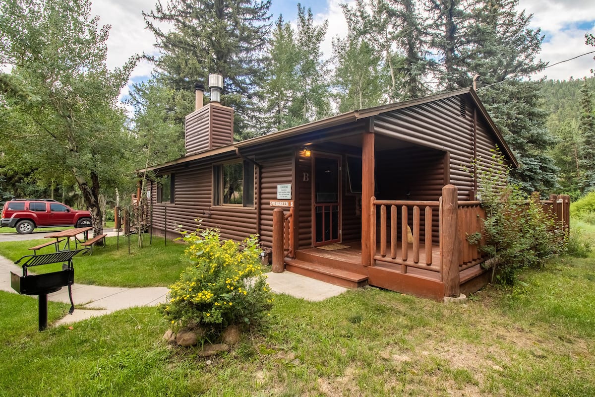 A rustic log cabin with a wooden porch, a chimney, and a picnic table.