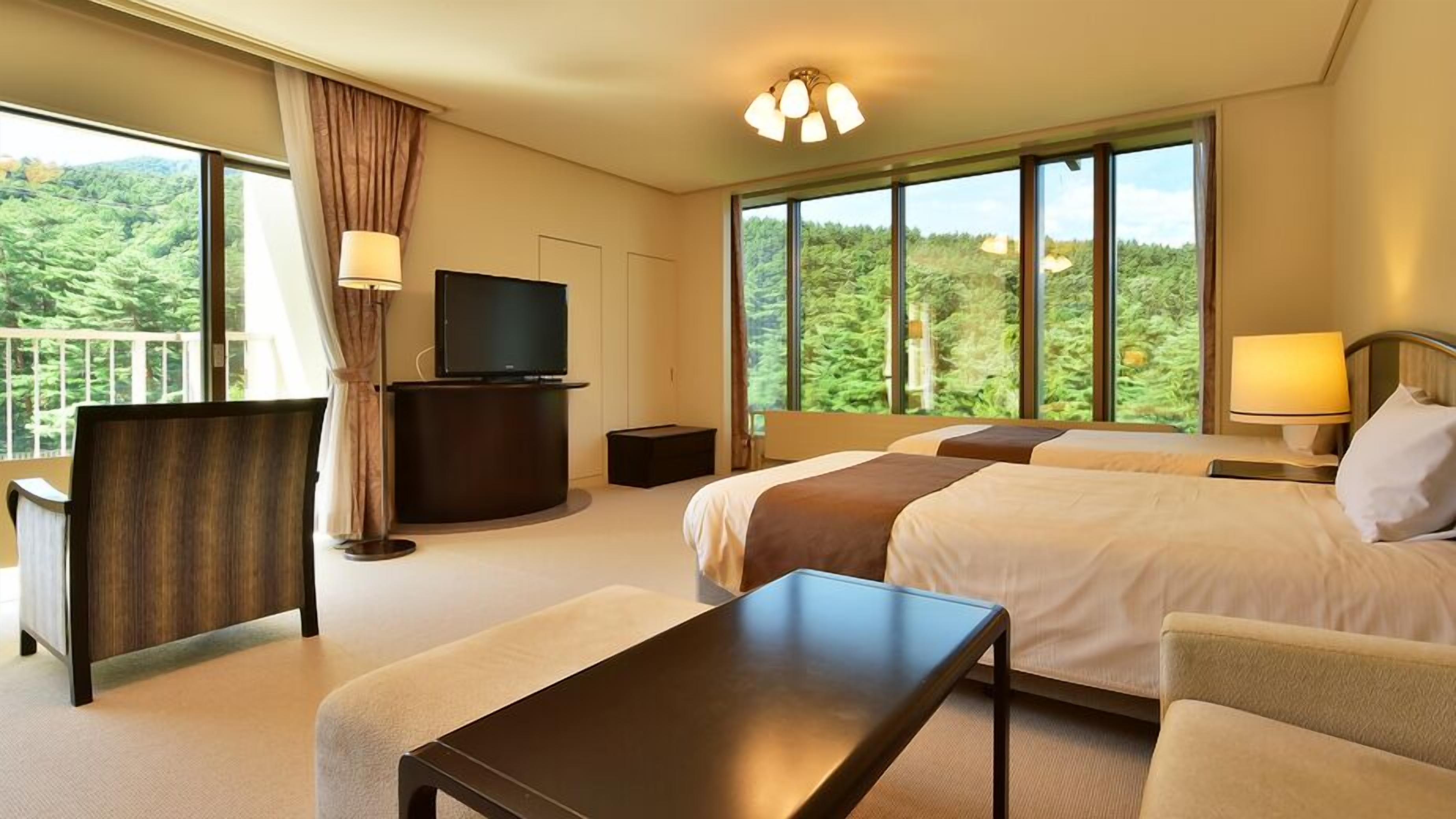 A hotel room with a bed, a desk, a TV, and large windows with a forest view.
