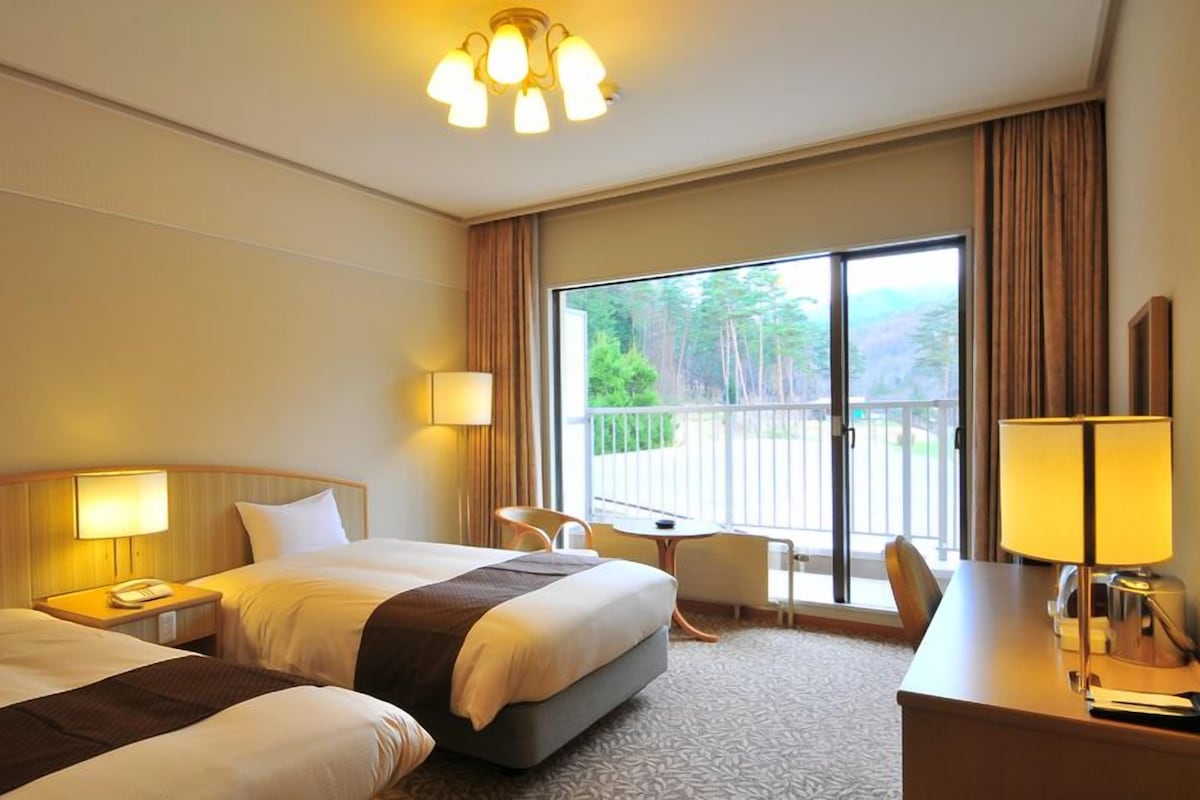 Habitación de hotel con dos camas, un escritorio, una silla, un balcón con vistas y un candelabro.