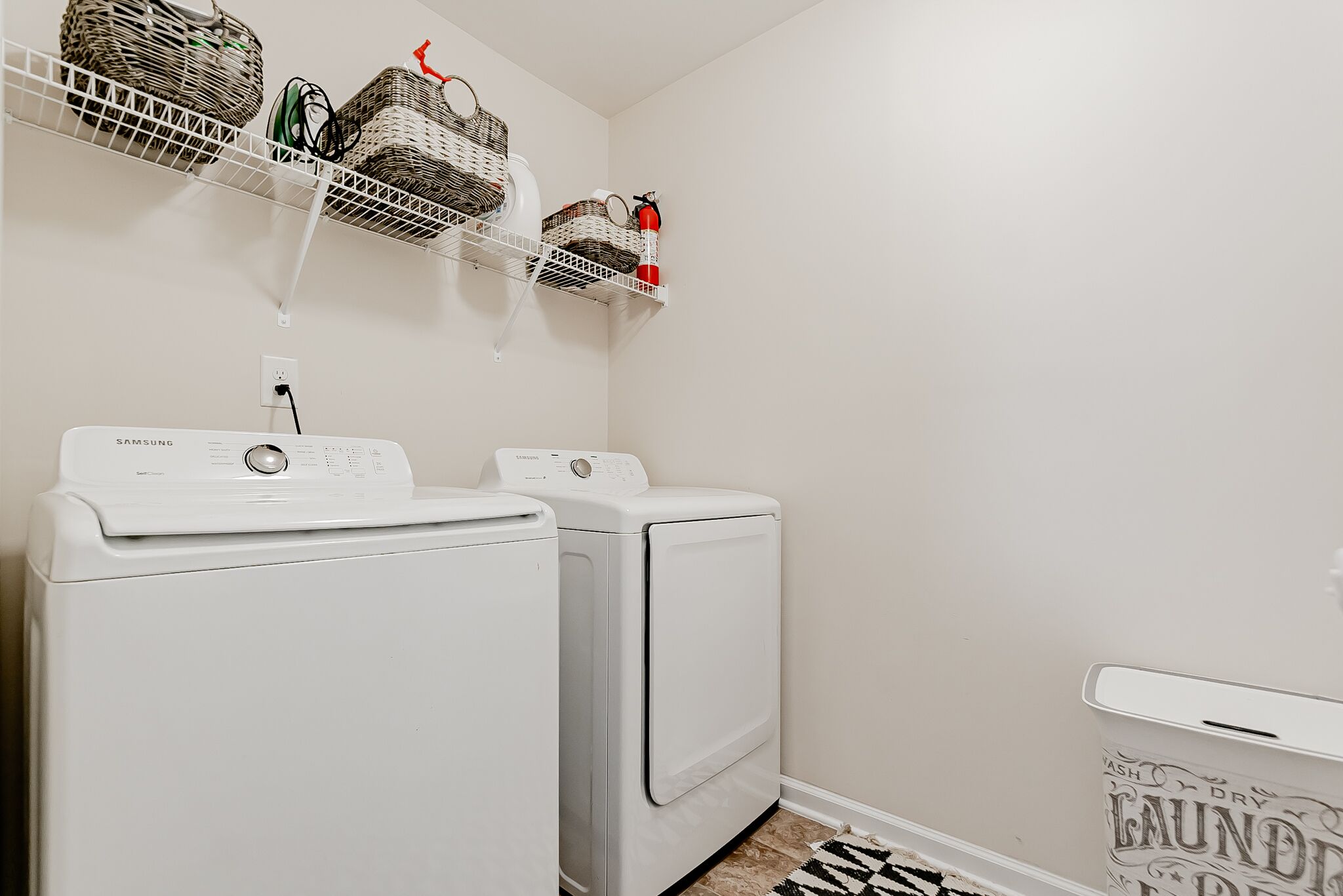 A laundry room with two white front-loading washers and dryers, a shelf with baskets and a fire extinguisher.