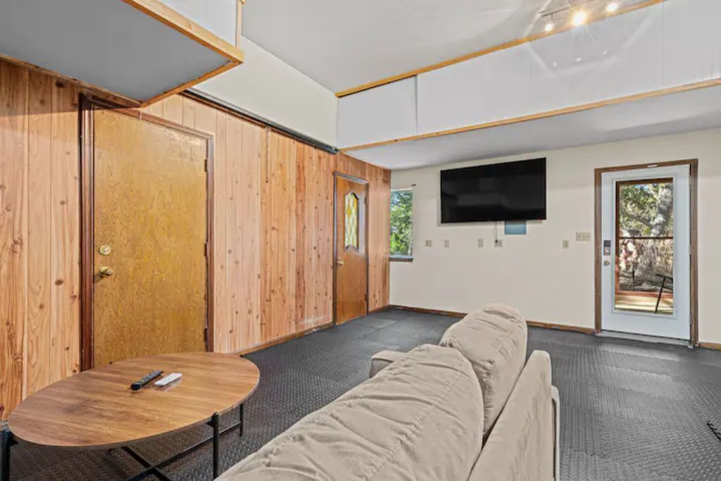 A room with a wooden door, a TV mounted on the wall, a sofa, and a small round table with a remote control.