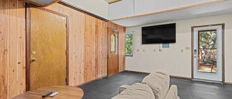 A room with a wooden door, a TV mounted on the wall, a sofa, and a small round table with a remote control.