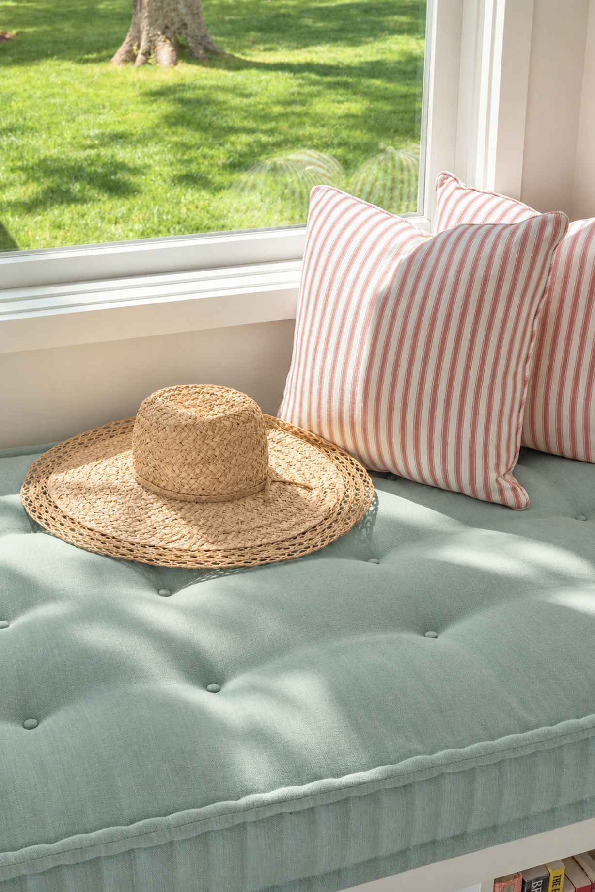 Asiento junto a la ventana con un sombrero tejido, almohadas a rayas y vista a un jardín verde.